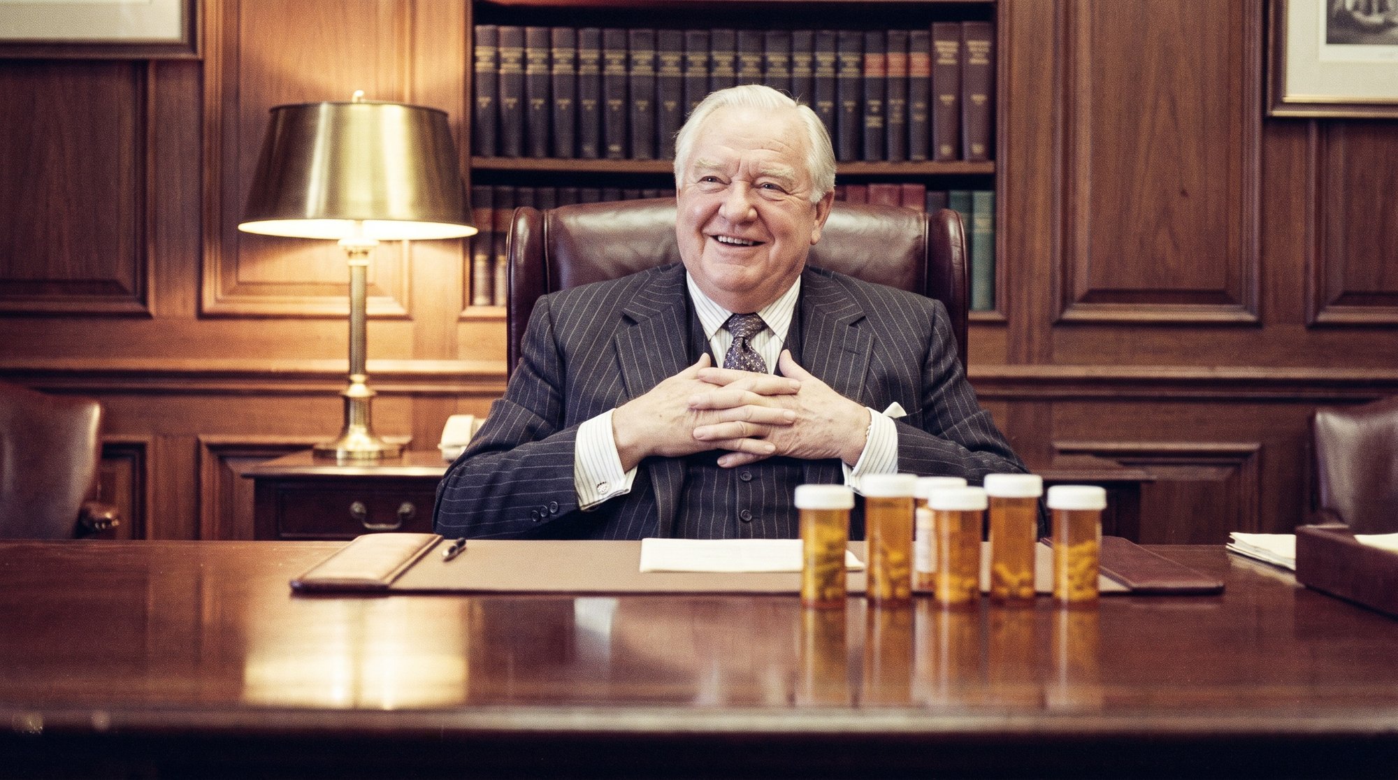 An elderly executive seated behind an enormous desk, smiling at the camera, with a row of prescription bottles lined up in front of him.